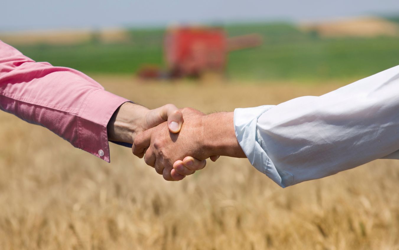 Close up of two businessmen shaking hands on the farmland. Combine harvester working in background